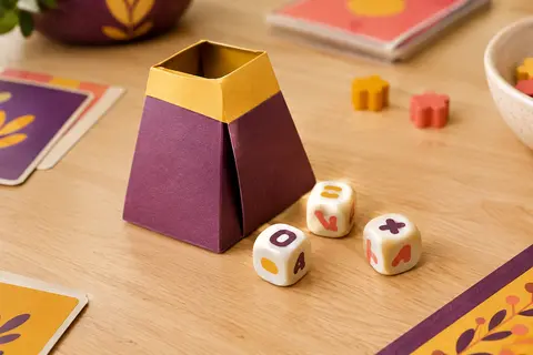 Shaker pliable en carton et trois dés de lettres posés sur une table de jeu en bois clair.