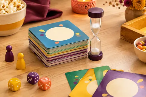 Jeu de cartes compact aux tons colorés avec un sablier, posé sur une table en bois clair.