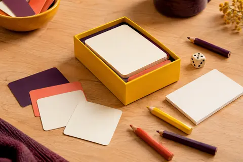 Boîte de jeu jaune, cartes, petit bloc de dessin et crayons disposés sur une table en bois clair.