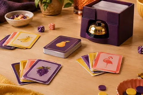 Jeu de cartes Pit avec étui rigide, sonnette intégrée et cartes multicolores sur une table en bois clair.