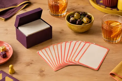 Petit étui en carton prune ouvert et cartes de mimes et devinettes étalées en éventail près de verres d’apéritif, sur une table en bois clair