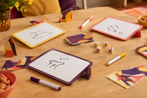 Ardoises effaçables, cartes et marqueurs du jeu de dessin réunis sur une table en bois clair.