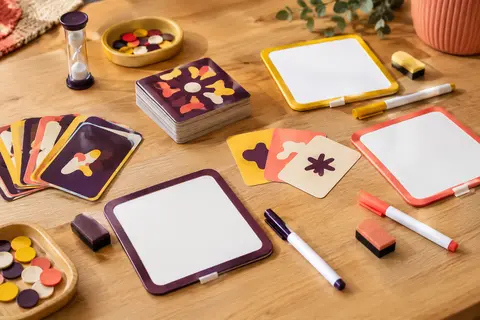 Cartes, tableaux effaçables et crayons d’un jeu coopératif de mots sur une table en bois clair.